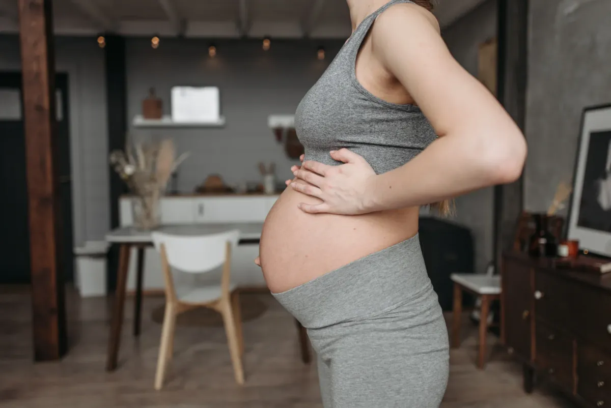 Femme enceinte debout dans son salon, une main posée sous son ventre arrondi