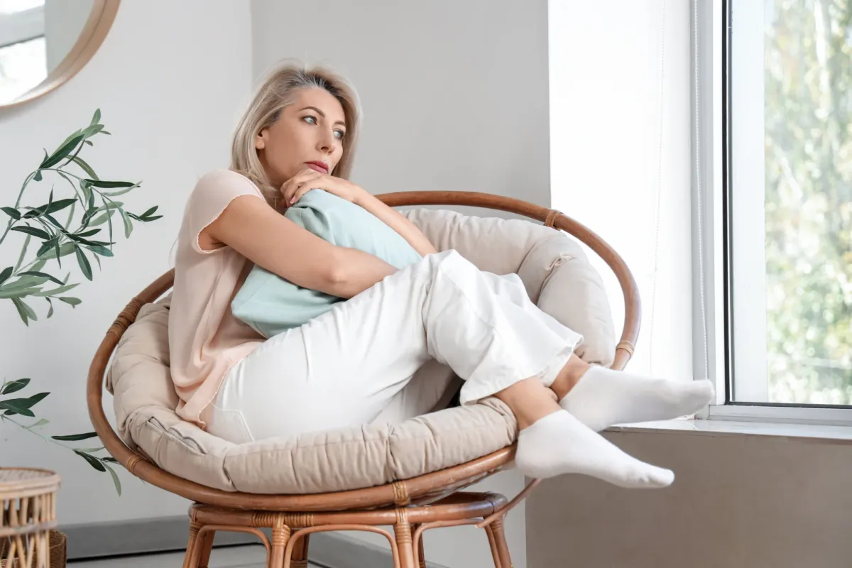Femme d'une cinquantaine d'années assise près d'une fenêtre, songeuse, un coussin serré contre elle