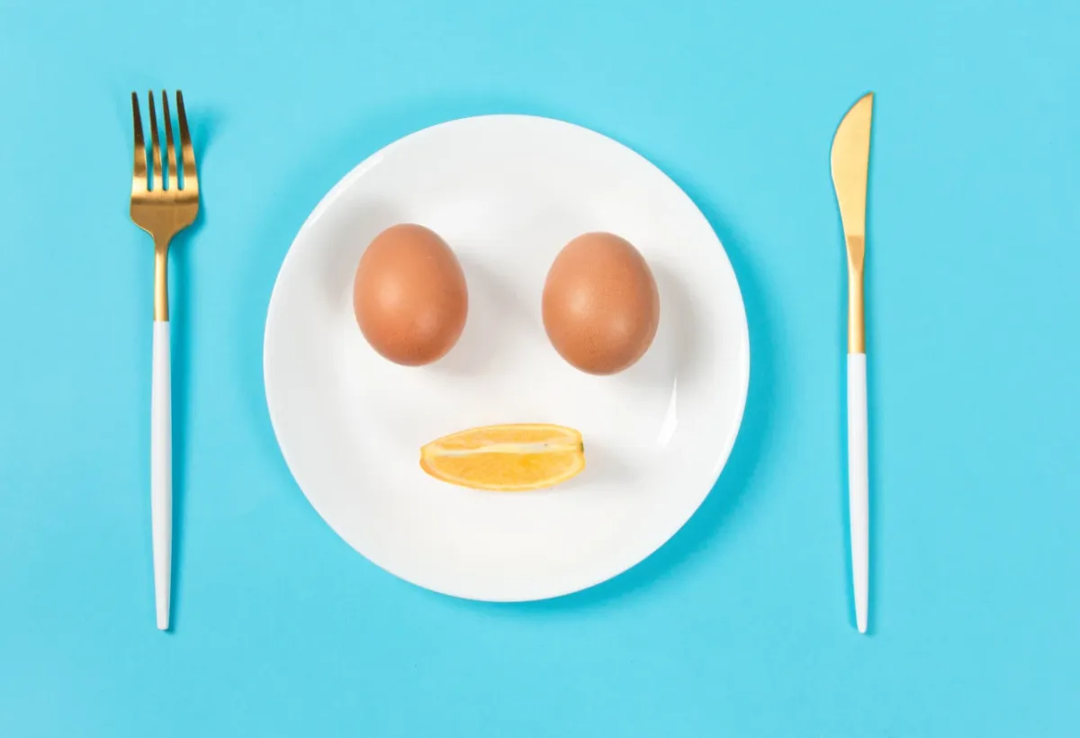 Assiette blanche composée en visage souriant avec deux œufs et un quartier d'orange, couverts dorés sur fond bleu