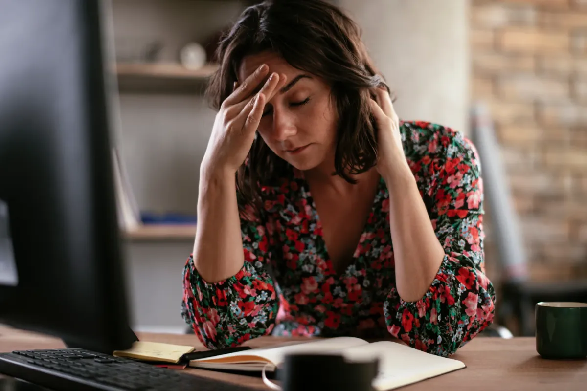Femme assise à son bureau devant son écran, les mains sur les tempes, les yeux fermés