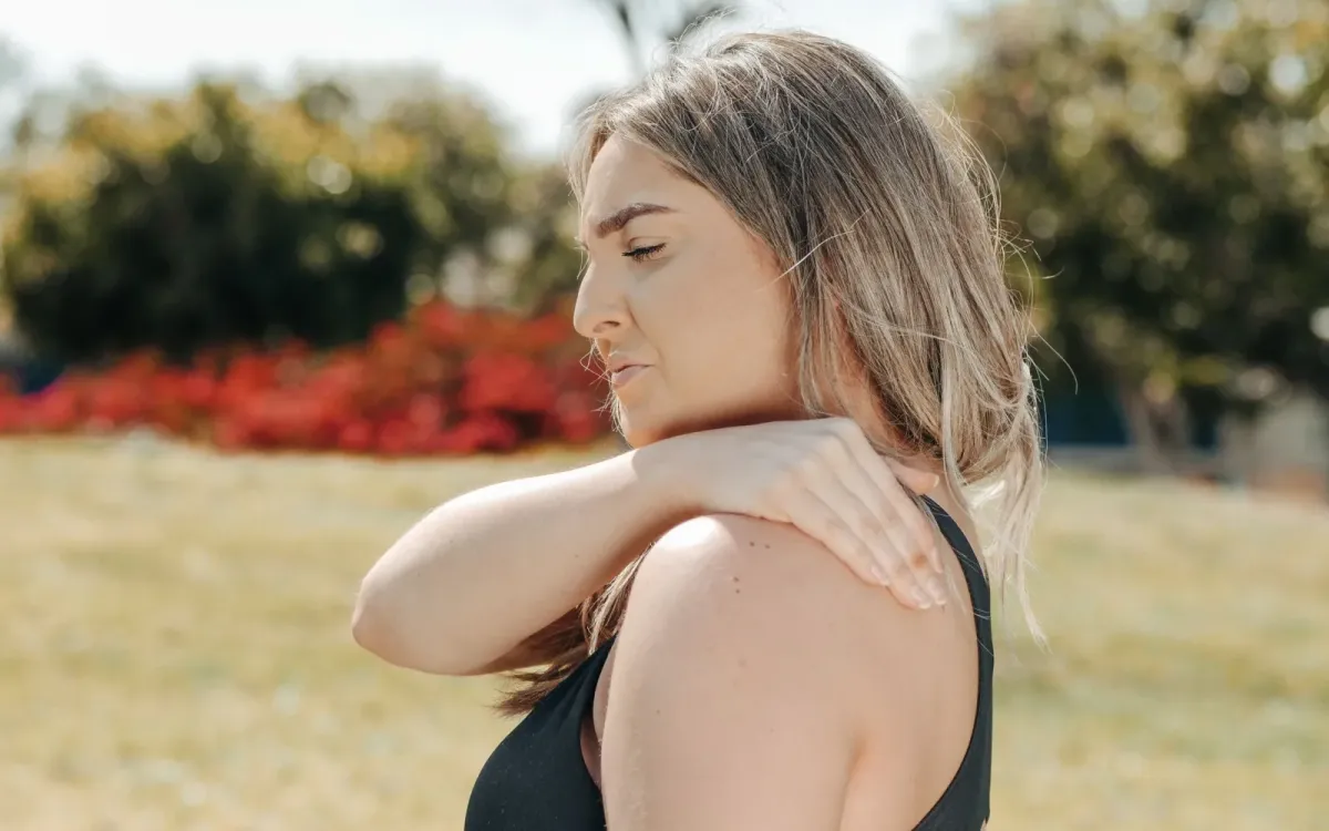 Femme dehors, le visage crispé, la main posée sur son épaule douloureuse