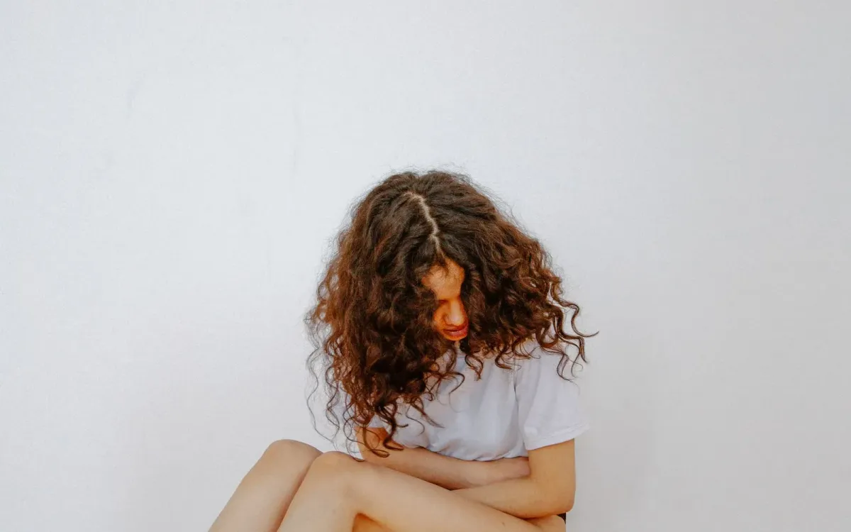 Femme assise au sol, genoux repliés contre elle, cheveux bouclés retombant sur son visage