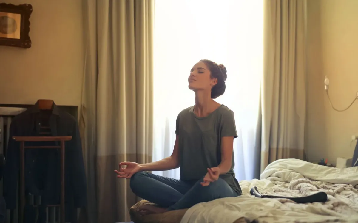 Femme assise sur son lit, les yeux fermés, dans une chambre calme