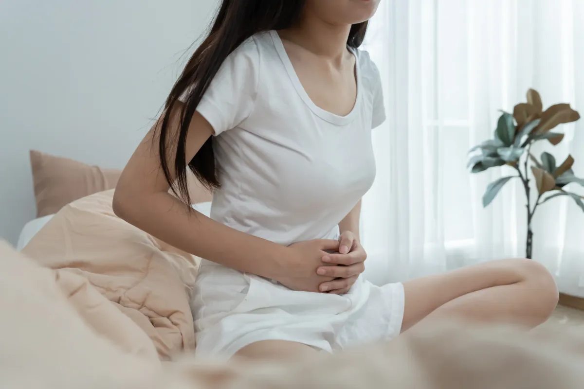 Jeune femme assise au bord de son lit dans la lumière du matin, une main posée sur le ventre