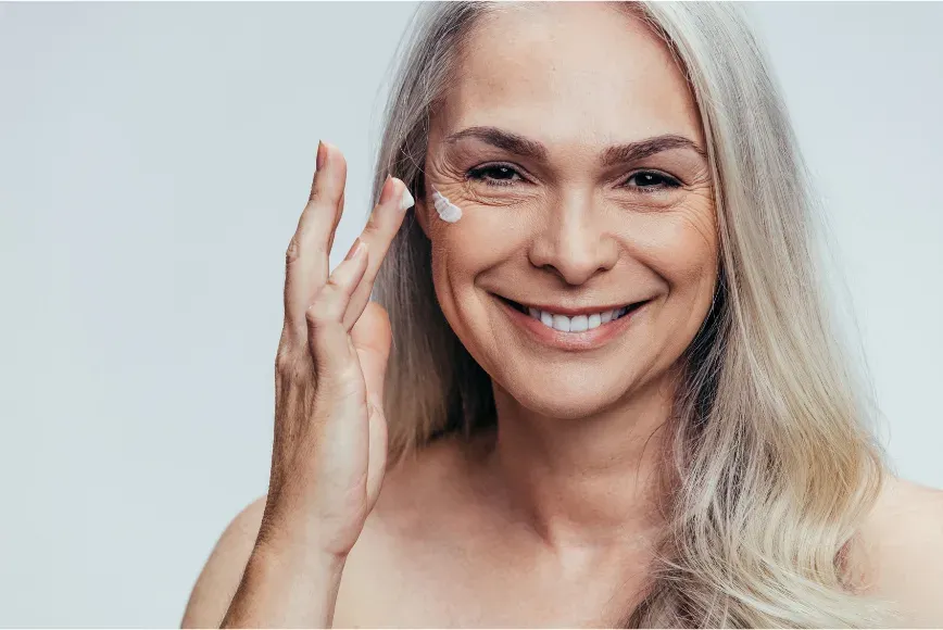 Femme aux cheveux gris appliquant une crème sur sa joue en souriant