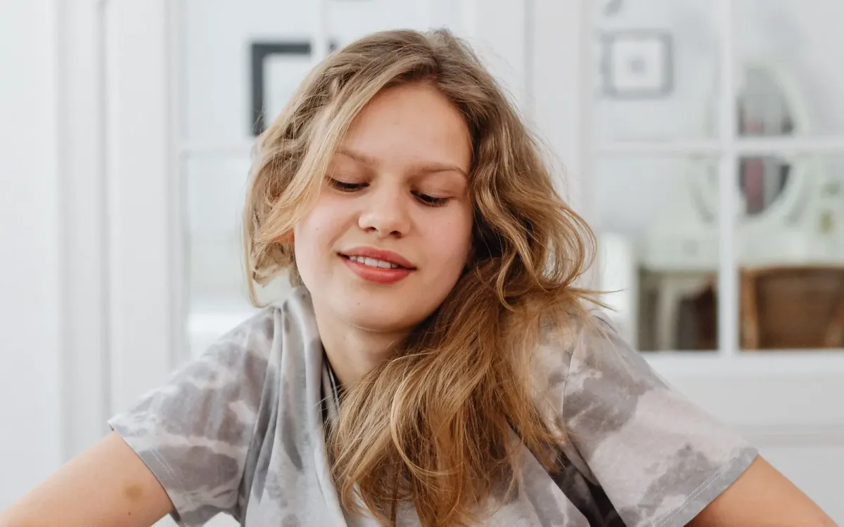Adolescente aux cheveux blonds ondulés, assise chez elle, le regard baissé et le sourire détendu