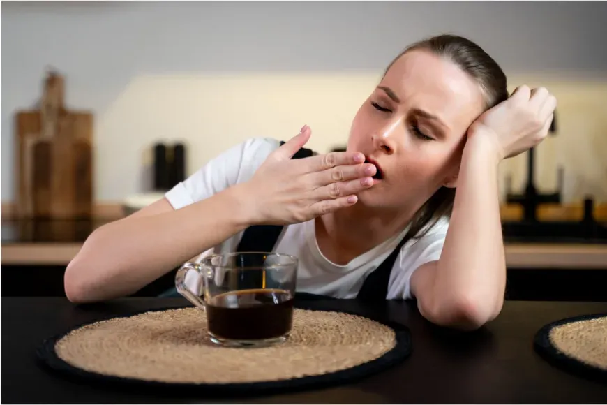 Femme qui bâille attablée devant une tasse de café le matin