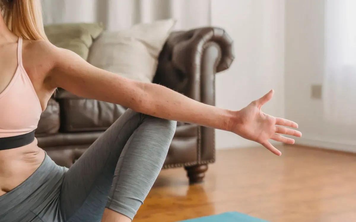 Femme en brassière rose assise sur un tapis dans son salon, bras tendu, en plein mouvement de gainage