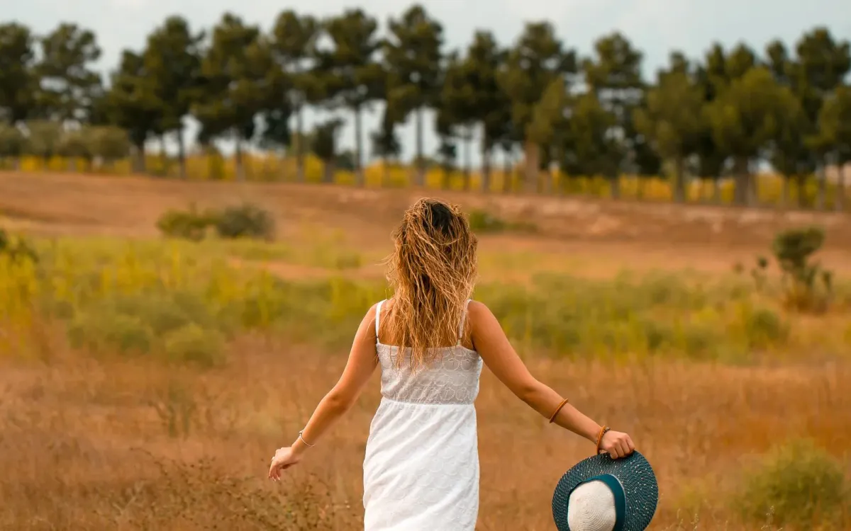 Femme en robe blanche marchant dans un champ doré, chapeau à la main, de dos