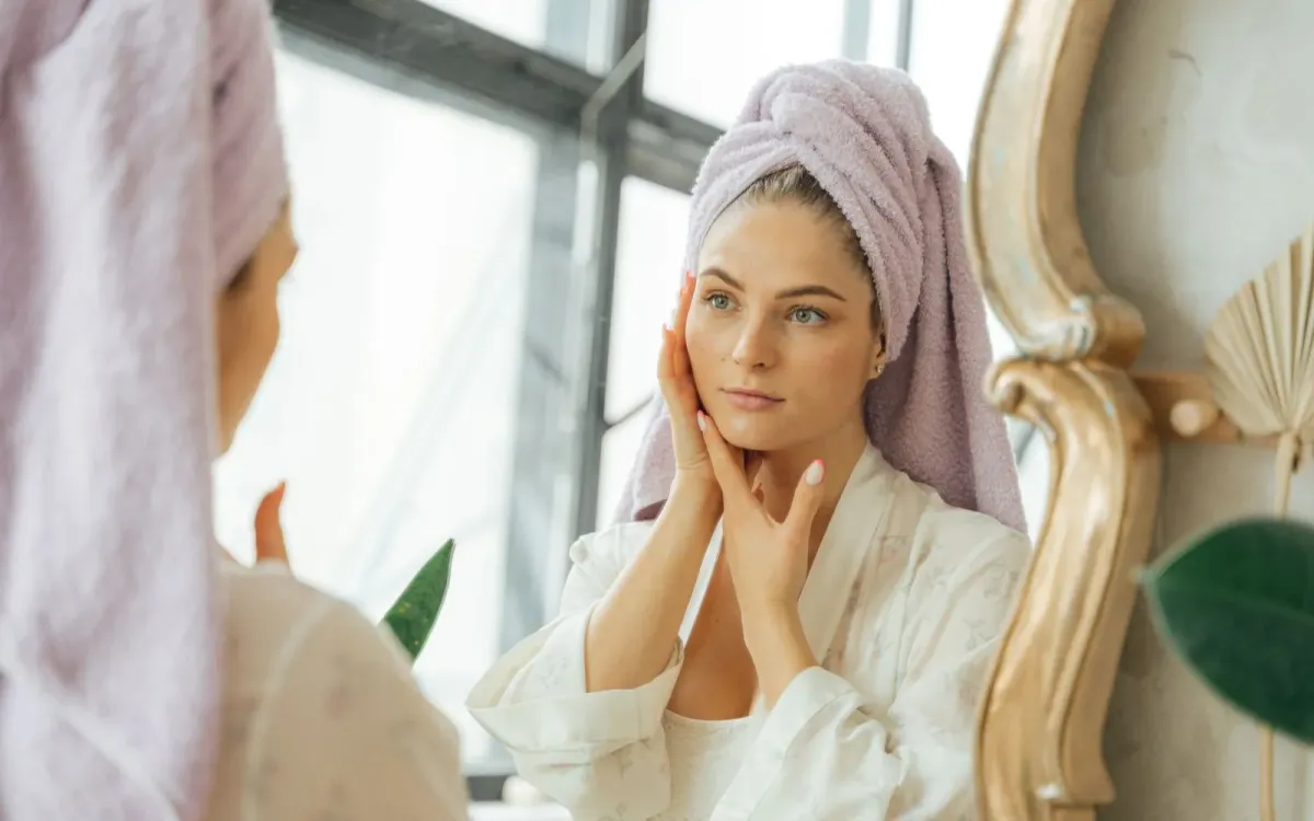 Femme en peignoir, une serviette sur la tête, qui touche son visage devant le miroir