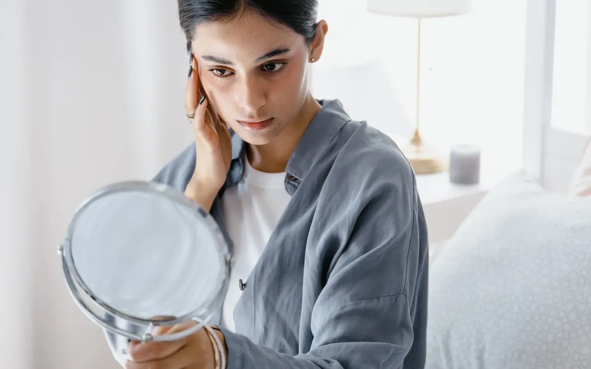 Femme assise sur son lit, qui touche son visage en se regardant dans un petit miroir