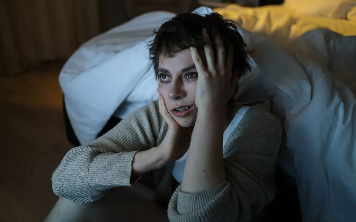 Femme assise au pied de son lit, la tête entre les mains, l'air préoccupé