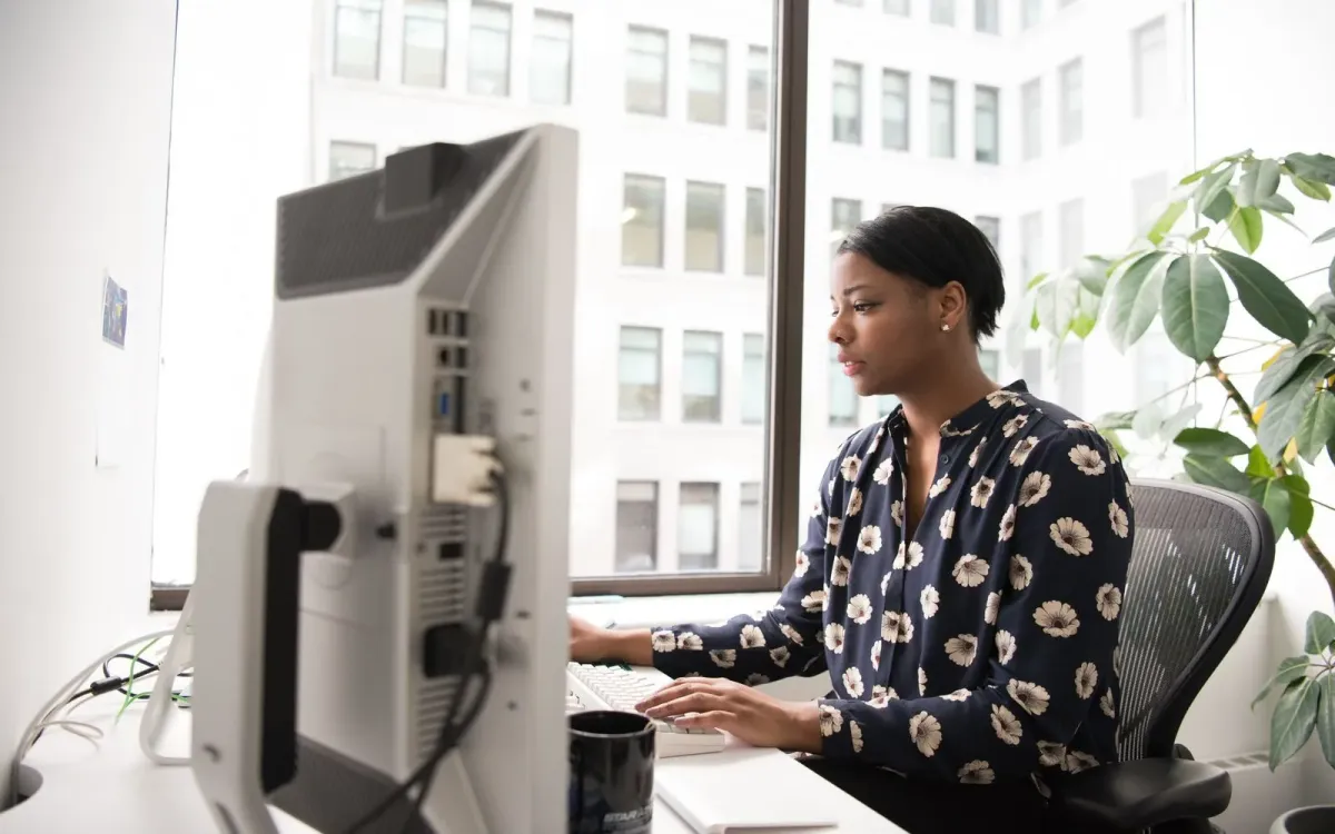 Femme en chemisier à fleurs qui tape sur un ordinateur de bureau, devant une grande fenêtre