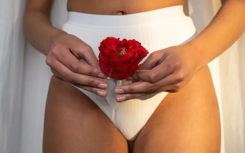 Femme en culotte blanche côtelée, tenant une cup menstruelle et une rose rouge devant son bas-ventre